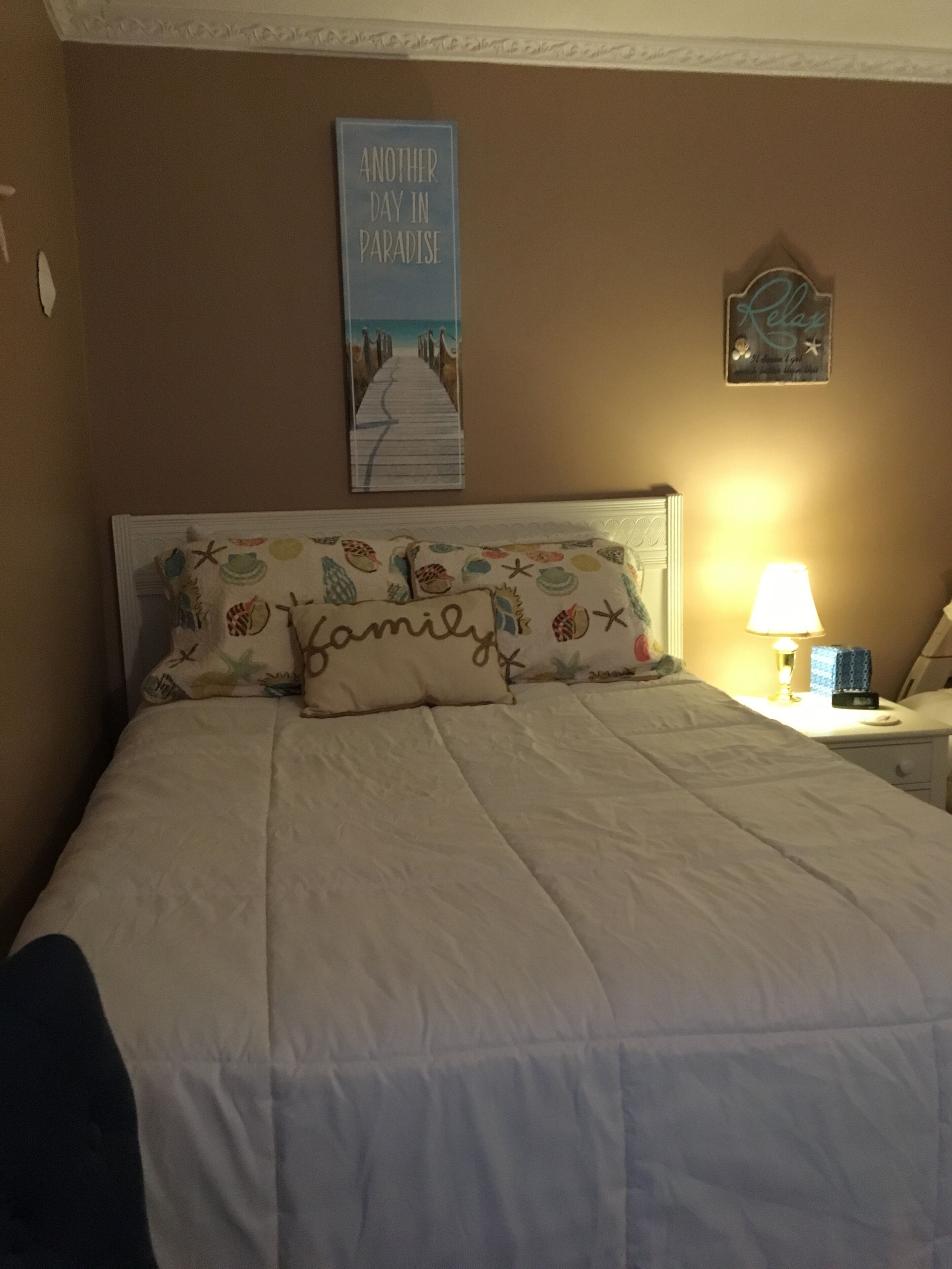 Une chambre avec un lit, un oreiller orné du mot « Famille », un mur avec une affiche sur laquelle on peut lire « Encore une journée au paradis », et une table de chevet avec une lampe.
