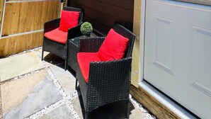 An outdoor seating area with wicker chairs and red cushions, a wooden fence, and a door with a window. - Luxury studio near IAH (Humble)