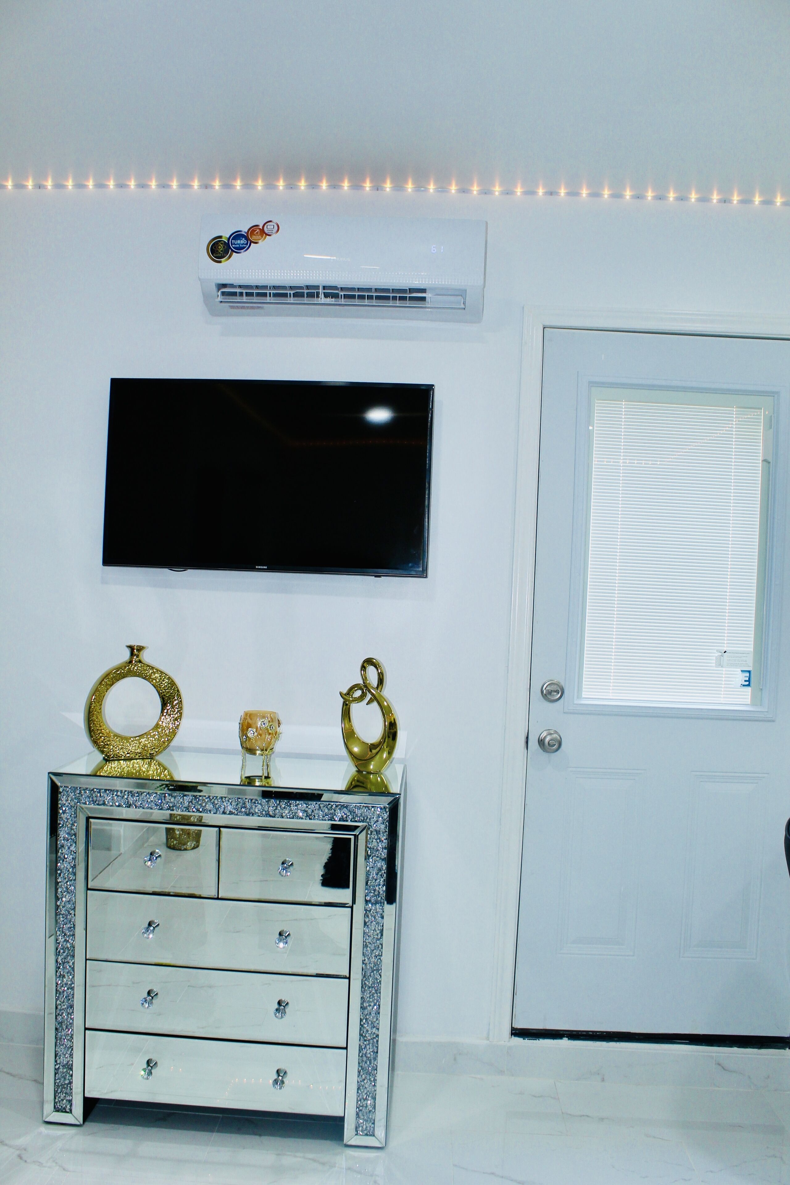 Una habitación blanca con un televisor montado en la pared, una puerta blanca con un ventanal y un tocador blanco con cajones.