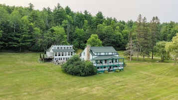Une grande maison à deux étages, dotée d’un toit végétalisé et d’une façade blanche, entourée d’une pelouse bien entretenue et d’une forêt dense.