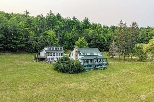Une grande maison à deux étages, dotée d’un toit végétalisé et d’une façade blanche, entourée d’une pelouse bien entretenue et d’une forêt dense.
