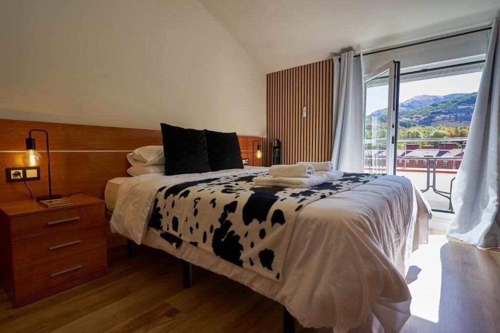 Un dormitorio con cabecera de madera, una cama con manta de estampado de vaca, una mesita de noche con lámpara, y vistas a un balcón con edificios y colinas.