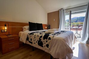 A bedroom with a wooden headboard, a bed with a cow print blanket, a bedside table with a lamp, and a view of a balcony with buildings and hills.