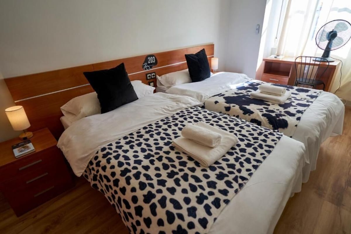 Habitación de hotel con dos camas, cada una con cabecera, mesita de noche con lámpara y un escritorio de madera con silla.