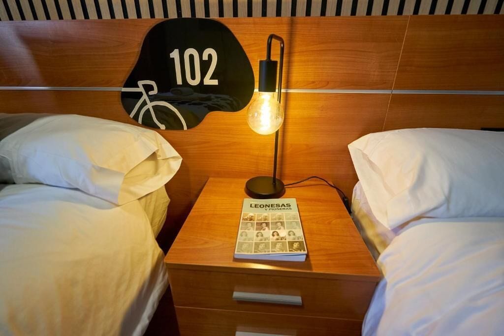 A hotel room with two beds, a nightstand with a lamp, and a wall-mounted sign with the number 102.