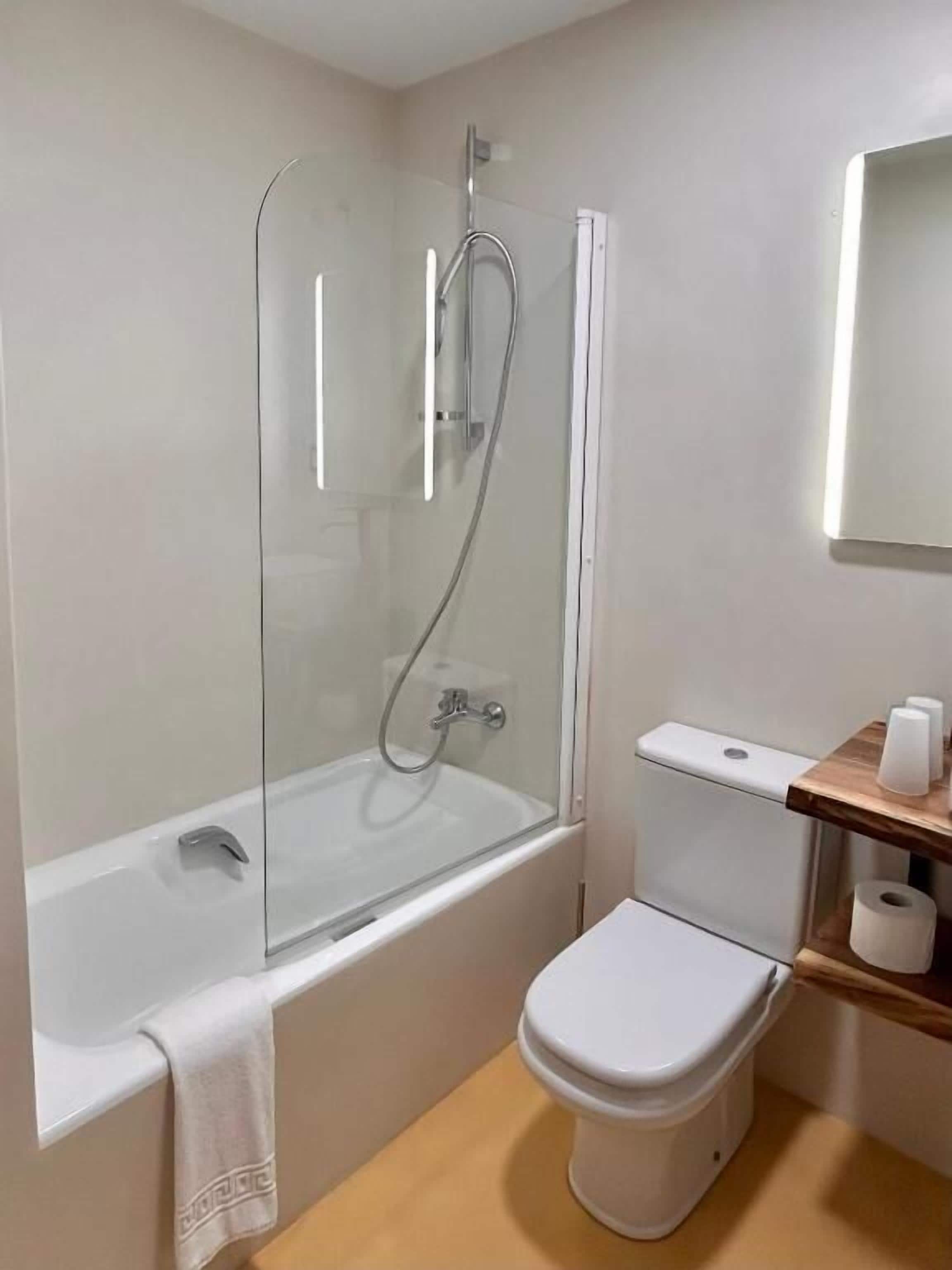 Un baño con ducha envuelta en vidrio, inodoro blanco y repisa de madera con papel higiénico.