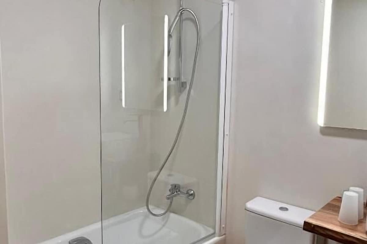 Un baño con ducha envuelta en vidrio, inodoro blanco y repisa de madera con papel higiénico.