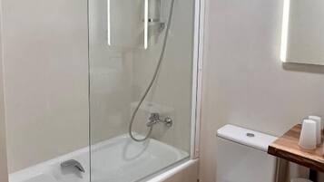 Un baño con ducha envuelta en vidrio, inodoro blanco y repisa de madera con papel higiénico.