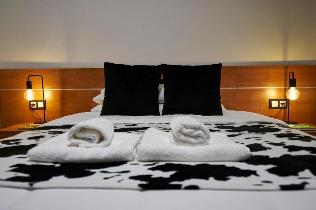 Habitación de hotel con cama, dos mesitas de noche, cabecera y dos toallas sobre la cama.