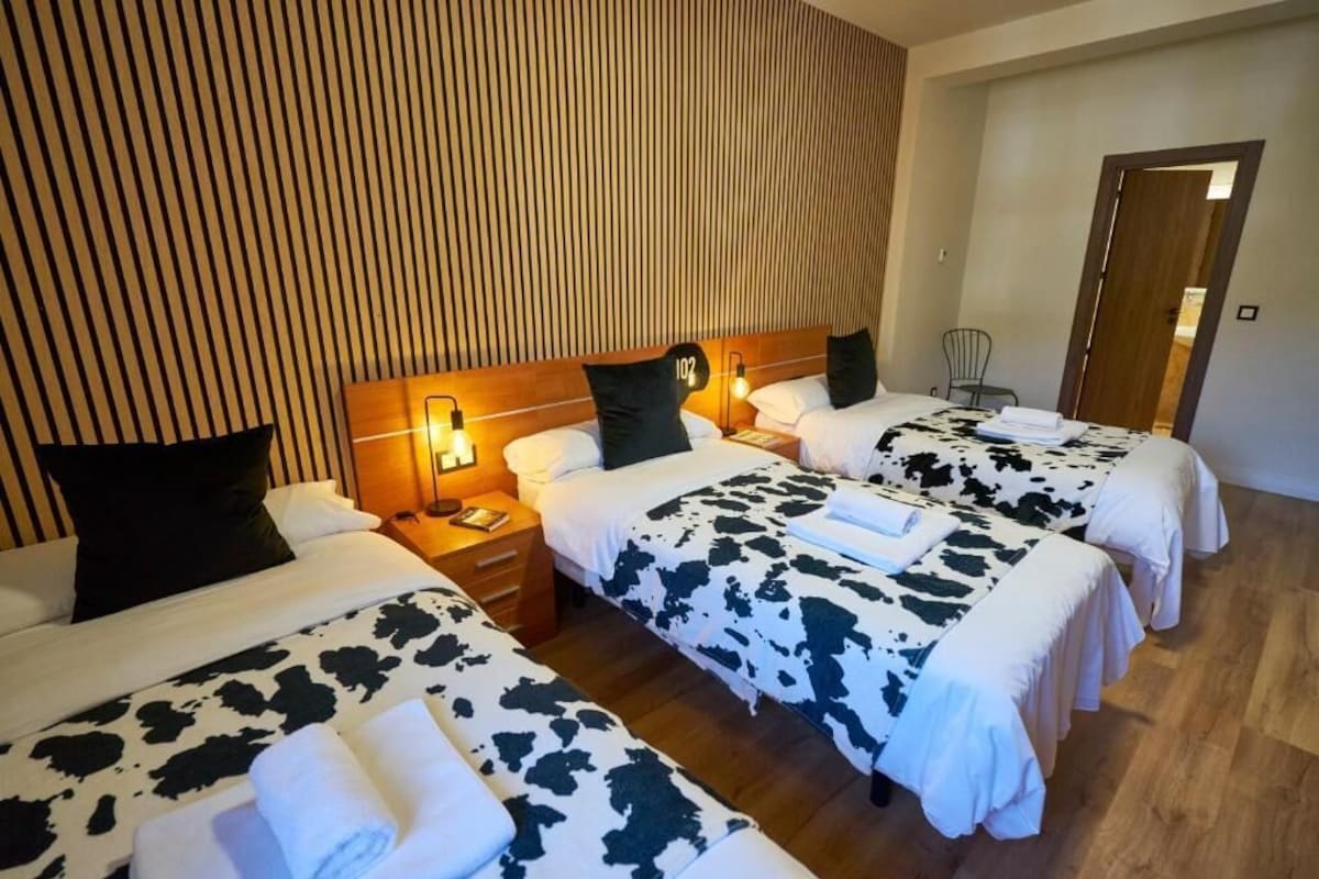 Habitación de hotel con tres camas, cada una con colcha de vaca, cabecero de madera y lámparas de noche.