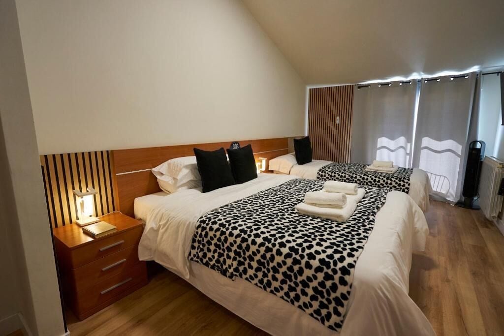Una habitación de hotel con dos camas, cada una con una colcha de estampado de leopardo, cabeceros de madera, mesitas de noche con lámparas y vistas al exterior a través de cortinas.