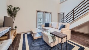 A modern living room with a sofa, coffee table, and TV stand. There is a staircase and a window with blinds. - Roomy 2B Loft Getaway | Sleeps 5 | Pool, Gym, Pkg, Pet Wash (Leander)
