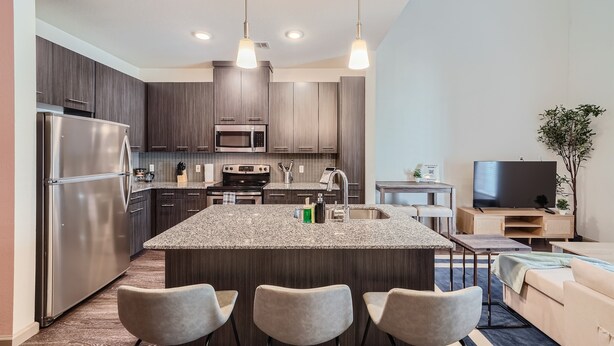 A modern kitchen with granite countertops, stainless steel appliances, and a central island with bar stools. - Roomy 2B Loft Getaway | Sleeps 5 | Pool, Gym, Pkg, Pet Wash (Leander)