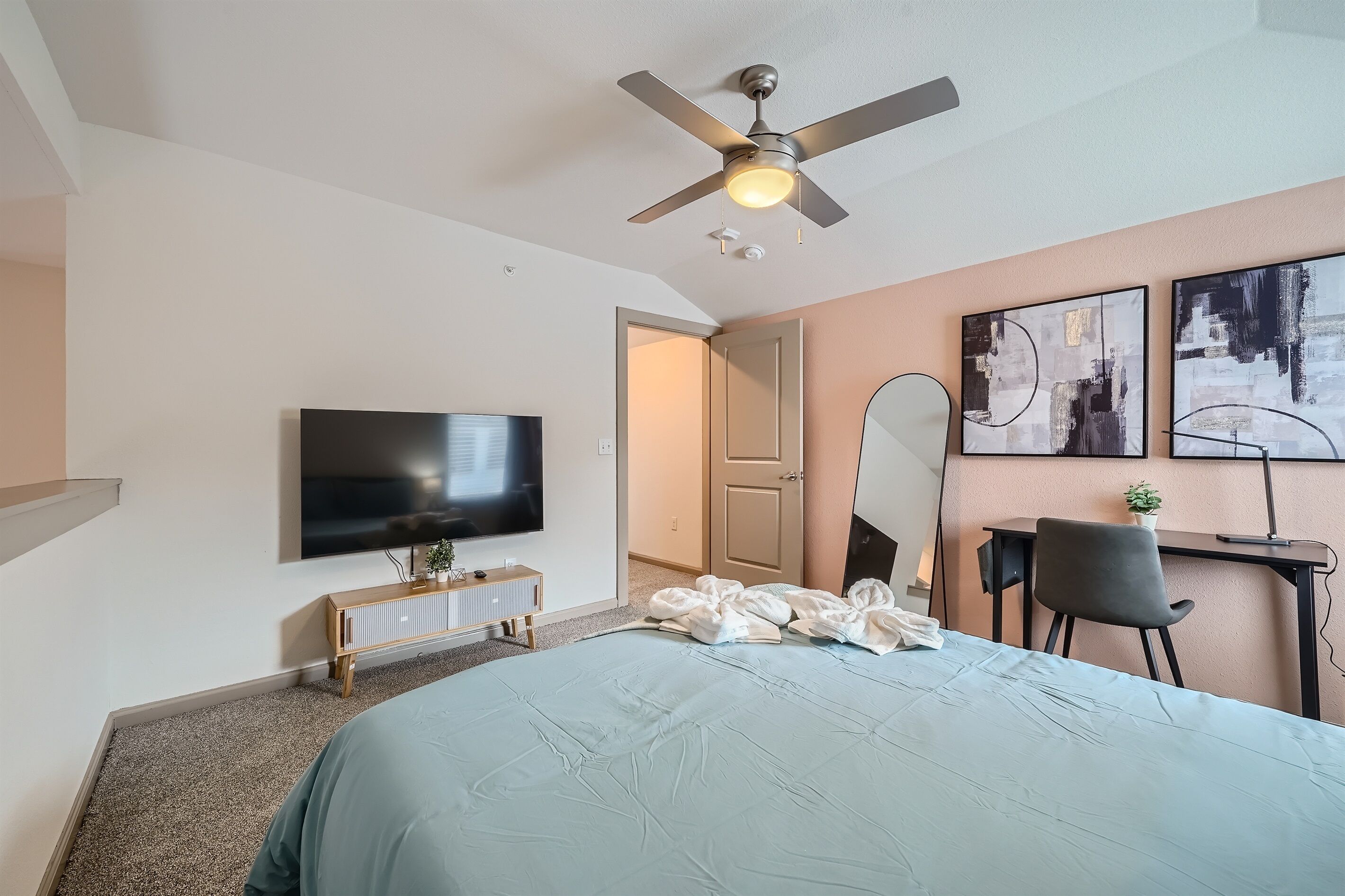 Una habitación de hotel moderna con una cama grande, ventilador de techo, televisor de pantalla plana, escritorio con silla y arte abstracto en la pared.