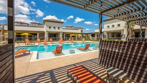 A pool area with lounge chairs and umbrellas, surrounded by buildings with a covered patio. - Roomy 2B Loft Getaway | Sleeps 5 | Pool, Gym, Pkg, Pet Wash (Leander)