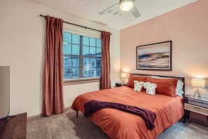 A bedroom with a large bed, a window with curtains, a nightstand with a lamp, and a painting on the wall. - Serene 3B Getaway | Pool, Gym, Parking, Pet Wash (Leander)