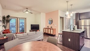 Ein modernes Wohnzimmer mit Sofa, Fernseher und Essbereich. Es gibt eine Küche mit Kühlschrank und einer kleinen Kochinsel mit Spüle.