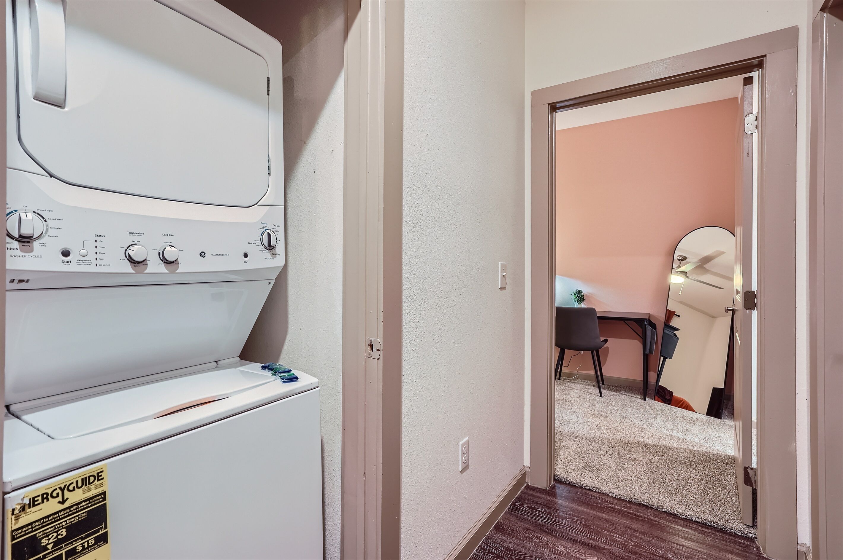 A laundry room with a front-loading washer and dryer, a door leading to a room with a desk and chair, and a carpeted floor.