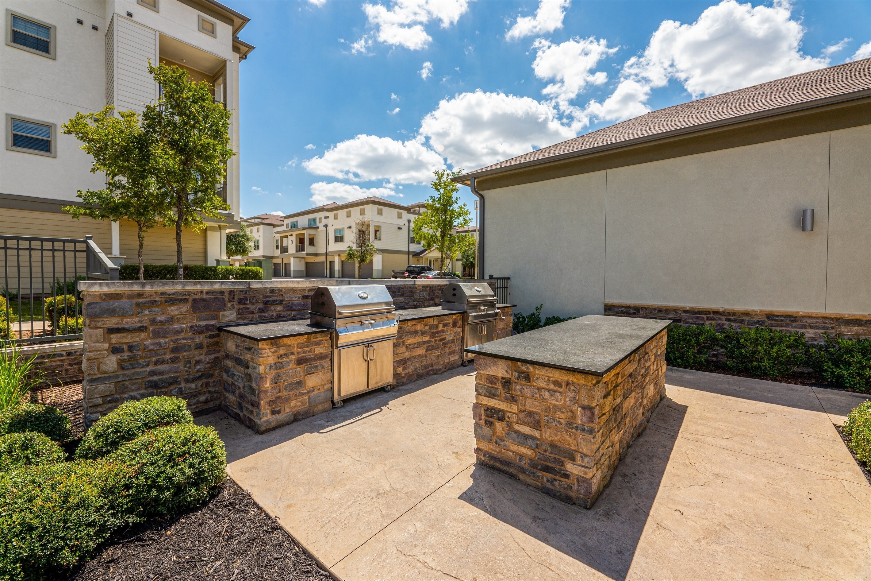 An outdoor patio with a stone barbecue setup, surrounded by a low stone wall and small shrubs.