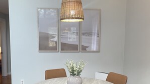 A round dining table with a textured white top, surrounded by four brown chairs. Above the table, there is a woven pendant light fixture. Three framed pictures are hung on the wall above the table. -  Cozy A-frame cabin just a short walk to town. (Ogunquit)