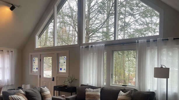 A cozy living room with a grey sofa set, a coffee table, and a large window with curtains. -  Cozy A-frame cabin just a short walk to town. (Ogunquit)