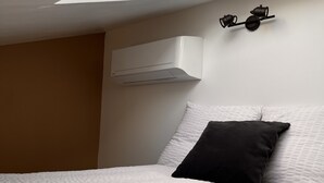 Une chambre avec un plafond en pente, un lit avec une literie blanche et un oreiller noir, et un climatiseur fixé au mur.