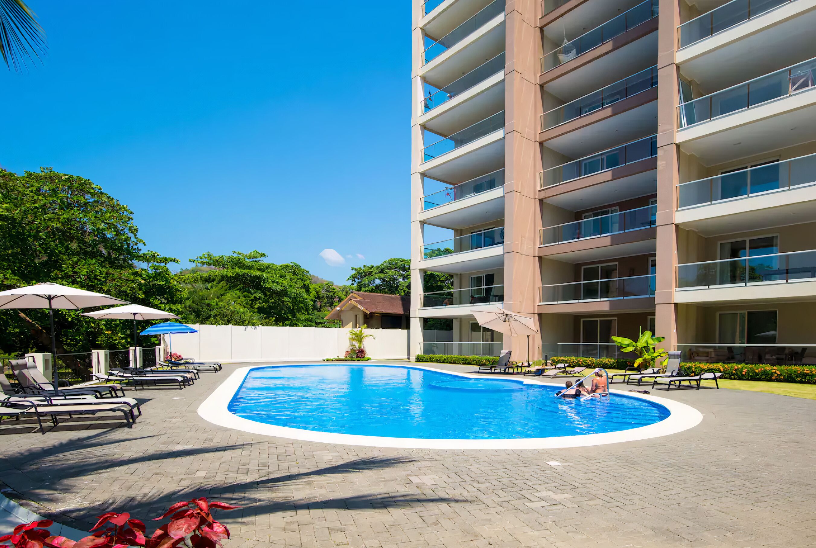 A swimming pool surrounded by lounge chairs and umbrellas, with a multi-story building in the background.