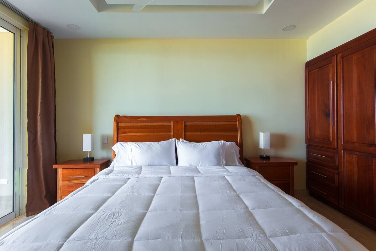 A neatly made bed with white linens in a room with wooden furniture and a large window.