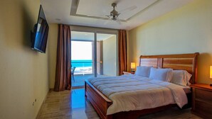 Hotelkamer met een bed, een aan de muur bevestigde televisie, een plafondventilator en uitzicht op de oceaan door een schuifglazen deur.