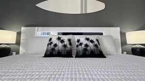 A modern hotel room with a large bed, two nightstands, a round mirror above the bed, and a window with blinds.