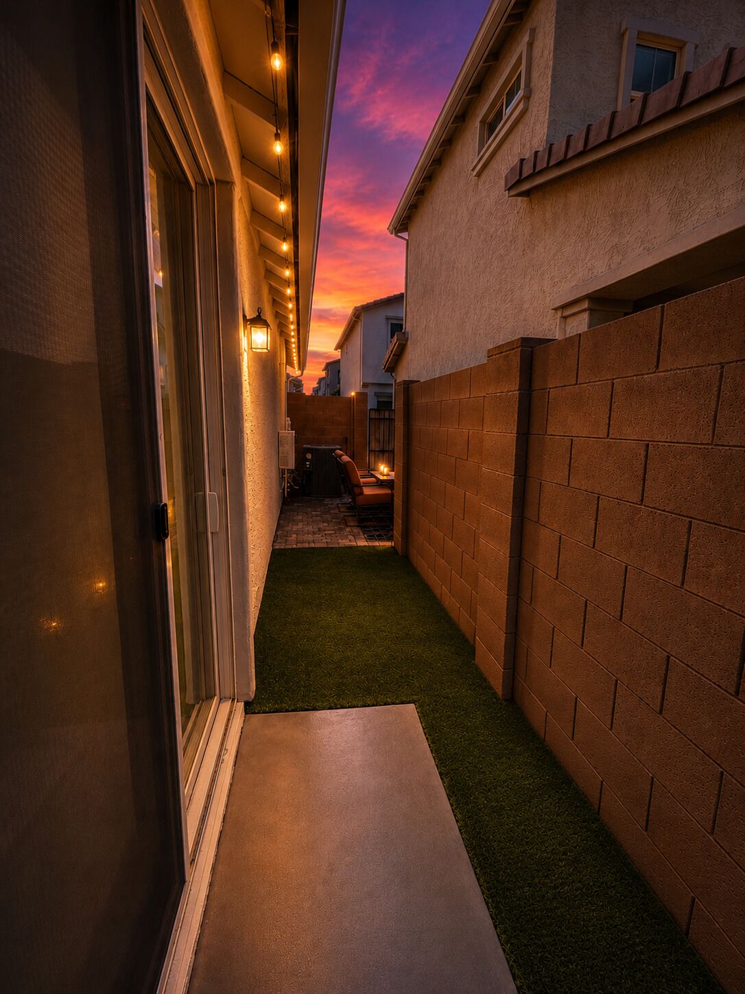 A narrow outdoor corridor with artificial lighting, a brick wall, and a small patch of artificial grass.