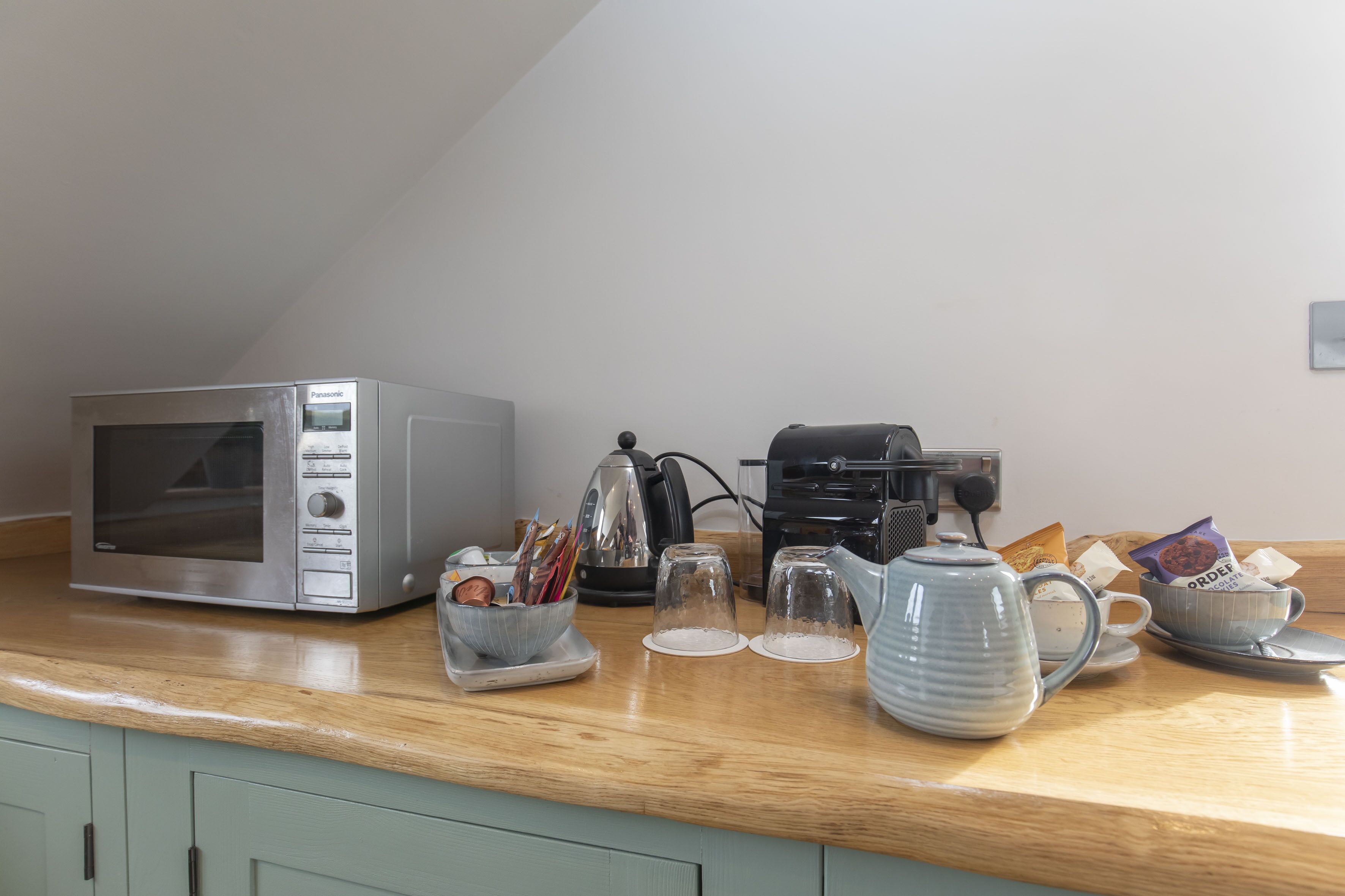 Een keuken met een magnetron, koffiezetapparaat, theepot en diverse keukengerei op het aanrecht.