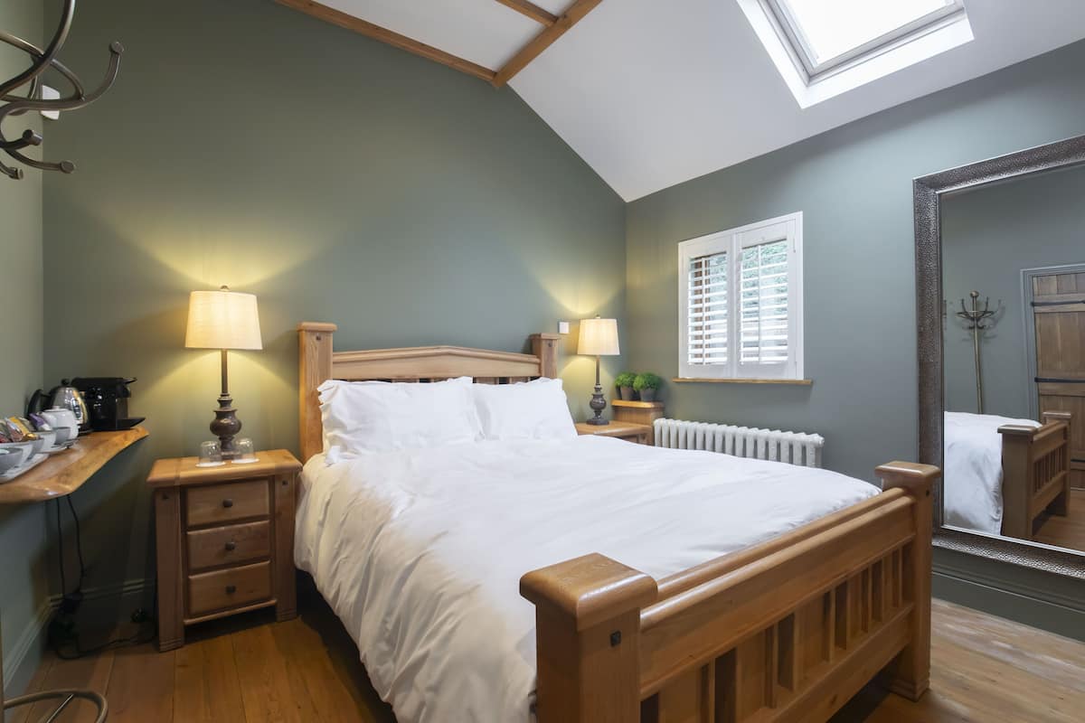A bedroom with a wooden bed, two bedside tables with lamps, a window with blinds, a radiator, and a mirror on the wall.