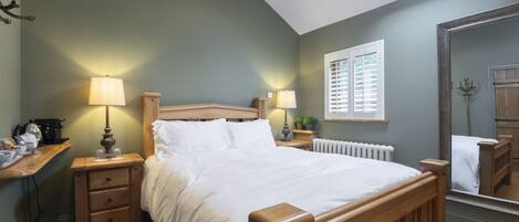 Quarto com cama de madeira, duas mesas de cabeceira com luminárias, janela com persianas, radiador e espelho na parede.
