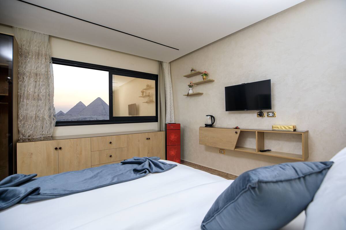A hotel room with a bed, a TV mounted on the wall, a window with a view of pyramids, a wooden cabinet, and a shelf with decorative items.