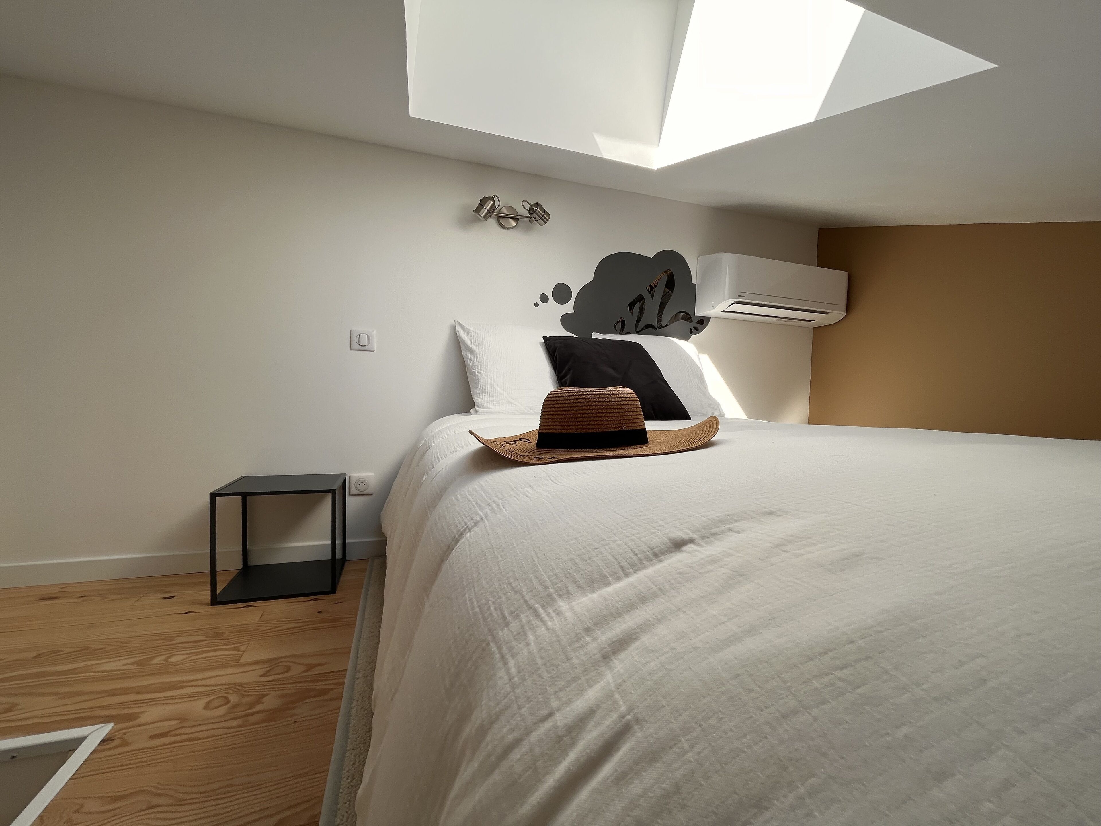 A bedroom with a skylight, a bed with white bedding, a wooden floor, a small bedside table, and a wall-mounted air conditioner.