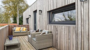 A modern outdoor deck with wicker furniture, a wooden deck, and a large window. - Luxury 6p. villa in Gaasterland near the woods and close to the (kite)beach. (Rijs)