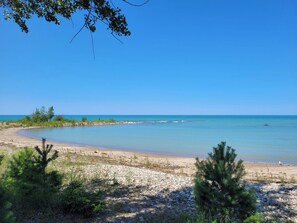 A beach with clear blue water, pebbles, and sparse vegetation.