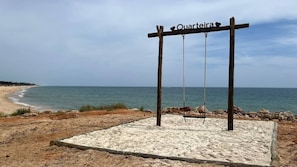 A wooden swing set with the word "Quartira" on it, positioned on a sandy area with a view of the beach and ocean.