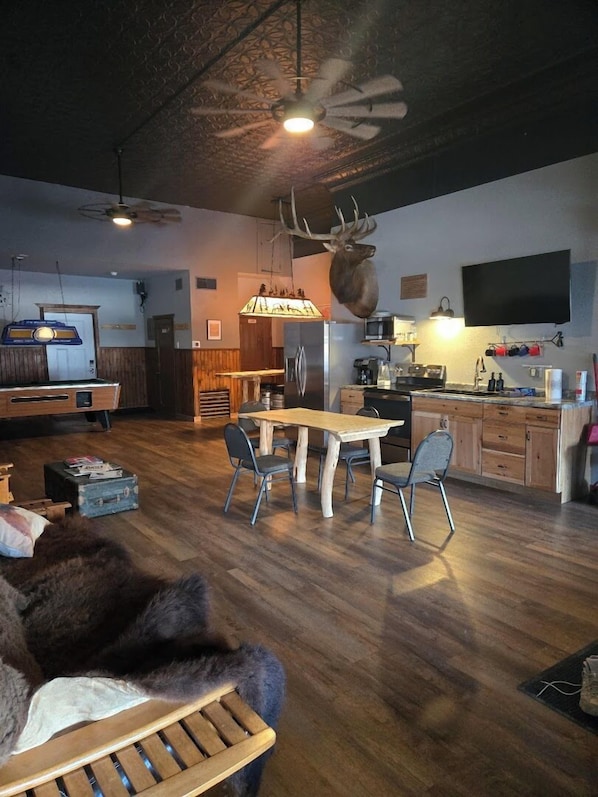 A spacious living area with a dining table, chairs, a mounted deer head, a television, and a kitchen area.