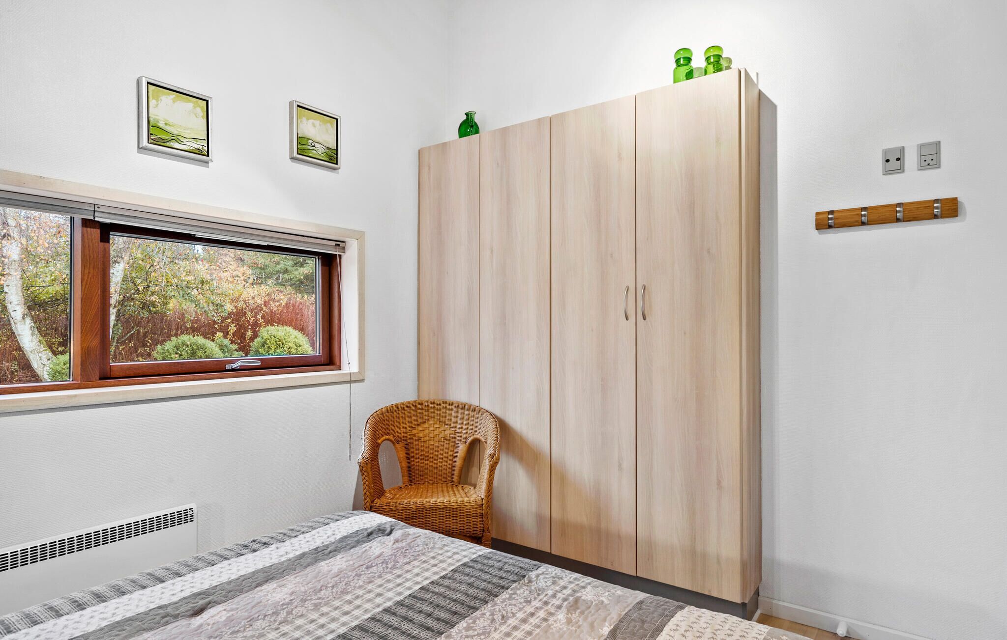 A bedroom with a large wardrobe, a wicker chair, a bed with a patterned bedspread, and two framed pictures on the wall.