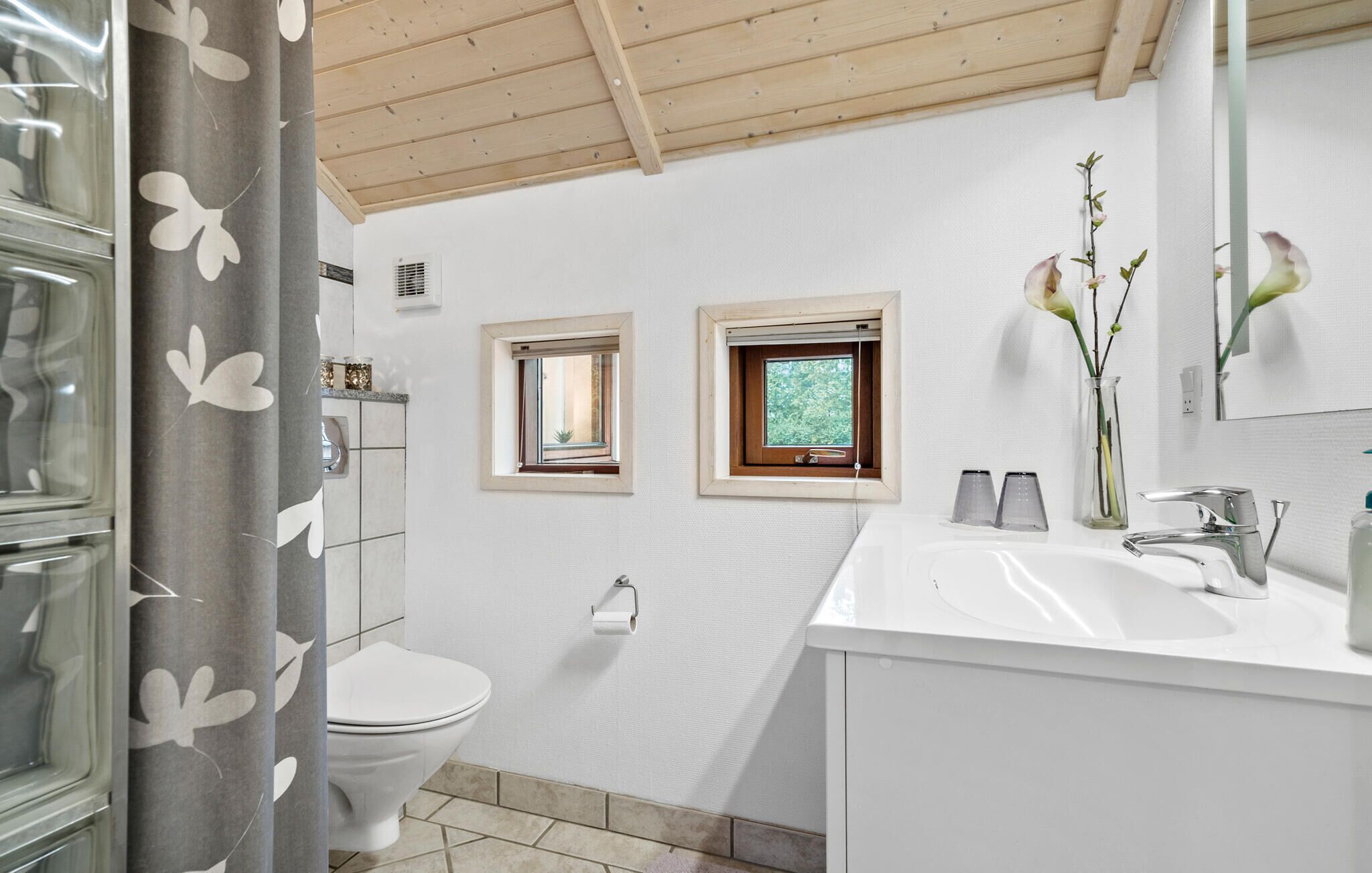 Une salle de bain comprenant des toilettes, un lavabo, un miroir et deux petites fenêtres.