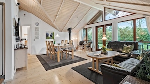 A spacious living area with a high ceiling, wooden beams, a dining table with chairs, a leather sofa, and large windows offering a view of the outdoors. - Awesome home in Ulfborg with kitchen (Ulfborg)