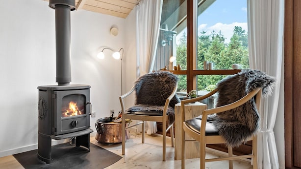 A cozy room with a wood-burning stove, two chairs with fur throws, a window with a view of trees, and a lamp on the wall. - Awesome home in Ulfborg with kitchen (Ulfborg)