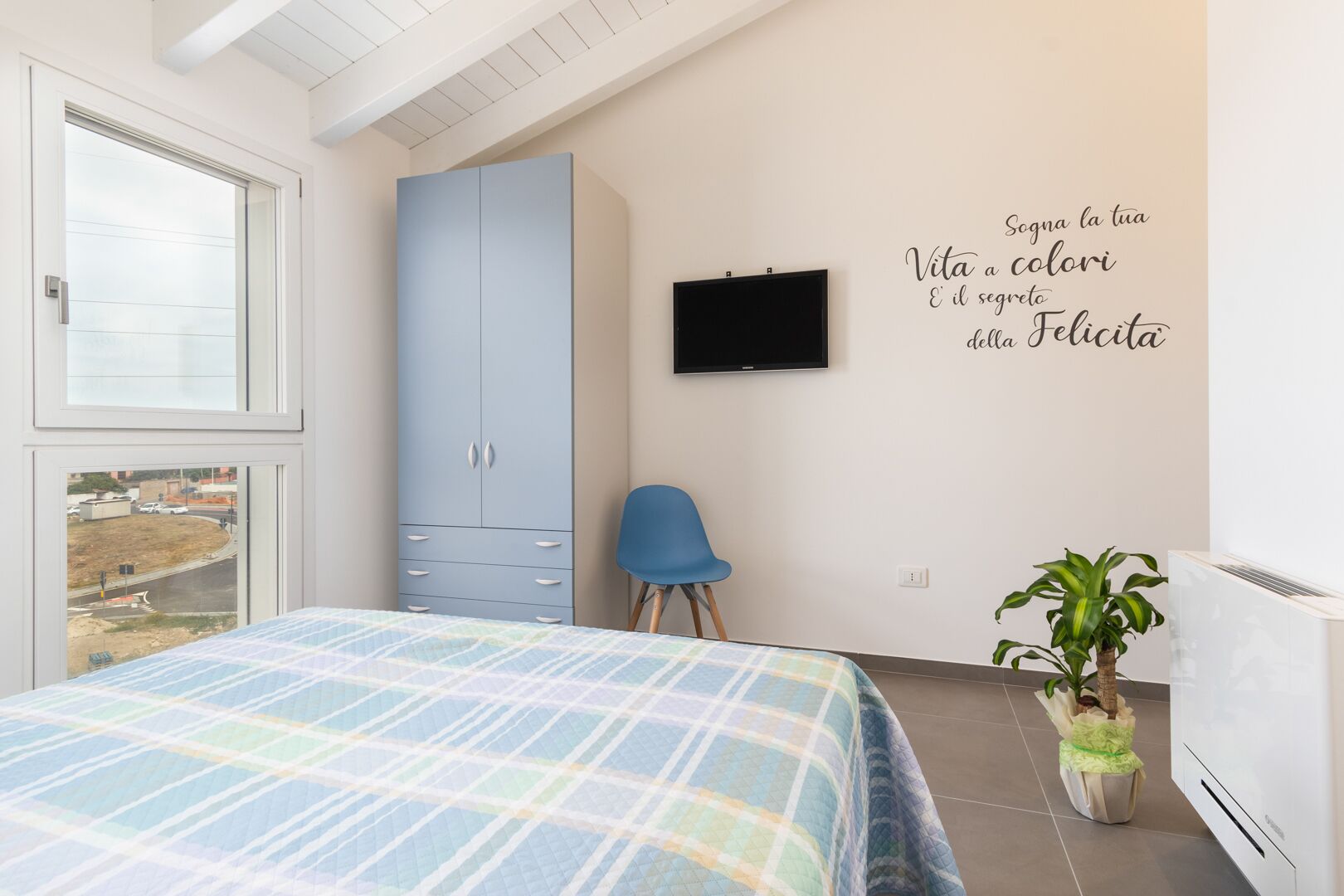 Un dormitorio con cama, armario, silla, televisor y una pared con una cita motivacional.