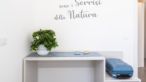 Una pared blanca con una frase decorativa en italiano, una planta en maceta, una pequeña mesa con dos tazas y una maleta azul sobre un banco blanco.