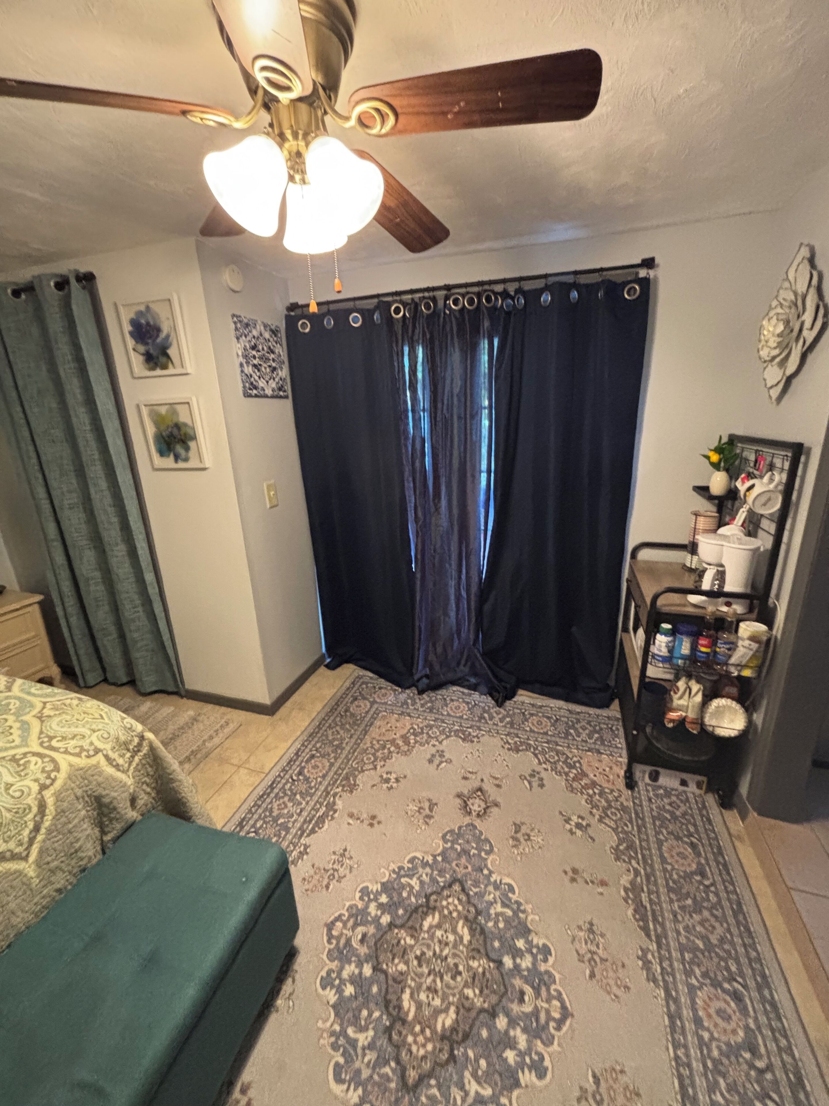 Ein Schlafzimmer mit Deckenventilator, einem Bett mit grüner Bettdecke, einem gemusterten Teppich, einem dunkelblauen Vorhang und einem kleinen Regal mit Gegenständen.