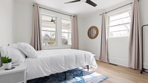 Une chambre à coucher avec un lit, un ventilateur de plafond, de grandes fenêtres avec des rideaux, une petite table de chevet avec une plante et un tapis à motifs.