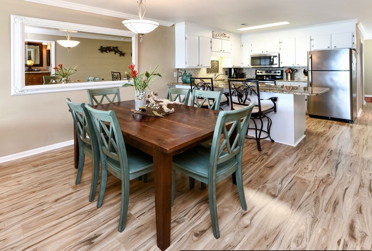 Un coin repas avec une table en bois et des chaises de couleur turquoise, une cuisine avec des meubles blancs et des appareils en acier inoxydable, et un plancher en bois.