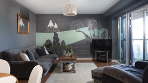 Ein modernes Wohnzimmer mit grauem Sofa, einem Couchtisch mit Büchern, einem Fernseher und einem Wandbild mit Berglandschaft.